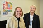 Nanna Hallikainen ja Sofia Åberg hymyilevät, taustalla näkyy kestävän kehityksen tavoitteita kuvaava juliste.