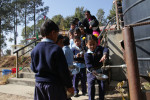 nepalilaisia lapsia koulun pihalla koulupuvuissa, tyttöjä ja poikia, huuhtelevat ruokakulhoja ennen ruokailua, vesitankki oikealla
