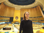 Hymyilevä nainen seisoo YK:n yleiskokousistuntosalin takaosassa. Taustalla kokoussalin penkkirivistöt ja puhujakoroke.