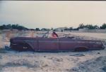 Vanha amerikanrauta hylättynä ja ilman renkaita Ambomaan hiekkaerämaalla, kuvassa auton ratissa poseeraa Helena Laukko