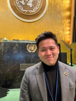 Nuorisodelegaatti Hung Ly hymyilee YK:n päämajan kokoustilassa. Takana näkyy YK:n logo seinällä ja puhujakorokkeen edessä.