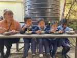 Kansanedustaja Aino-Kaisa Pekonen maistelee nepalilaista kouluruokaa vierellään neljä koululaista kouluasut yllään.