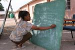 Nainen kirjoittaa liitutauluun kyykyssä. Taustalla koulualueen hiekkapiha aurinkoisena päivänä Botswanassa.