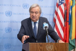 YK:n pääsihteeri puhujalavalla, mies, harmaat hiukset, puku ja solmio, taustalla lippuja