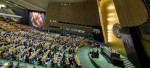 Ihmisiä istuu suuressa kokoussalissa, YK:n yleiskokoussali kuvattu ylhäältä päin